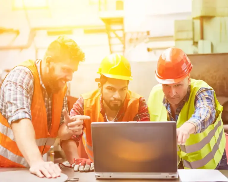 Three men wearing bright work vests and hard hats gathered around a laptop explaining and pointing to information.